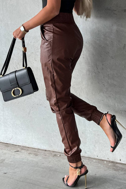 Person wearing brown leather jogger pants and black high-heeled shoes, holding a black handbag against a gray wall.