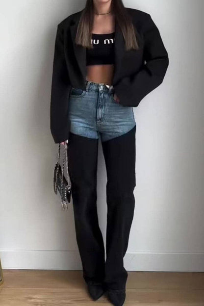 Person wearing a black jacket, black crop top, blue jeans, and black boots against a white wall.