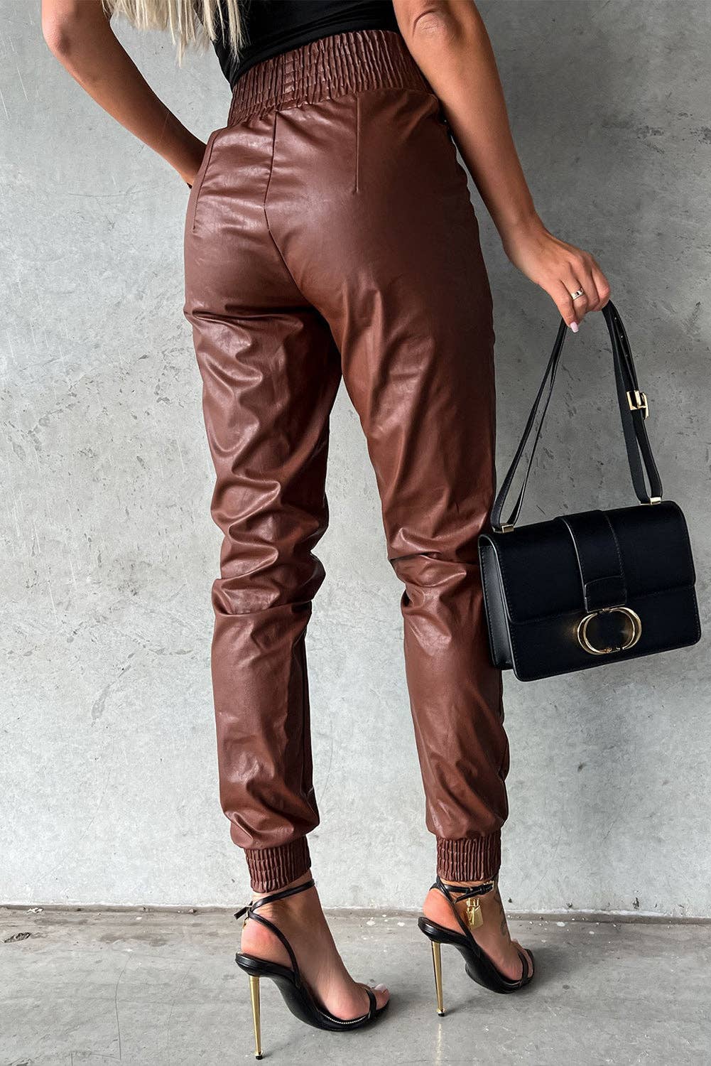 Person wearing brown leather jogger pants holding a black handbag against a gray background