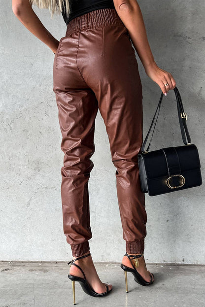 Person wearing brown leather jogger pants holding a black handbag against a gray background