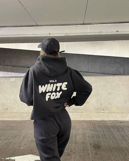 Person wearing a black hoodie with 'White Fox' text, standing against a modern architectural background.