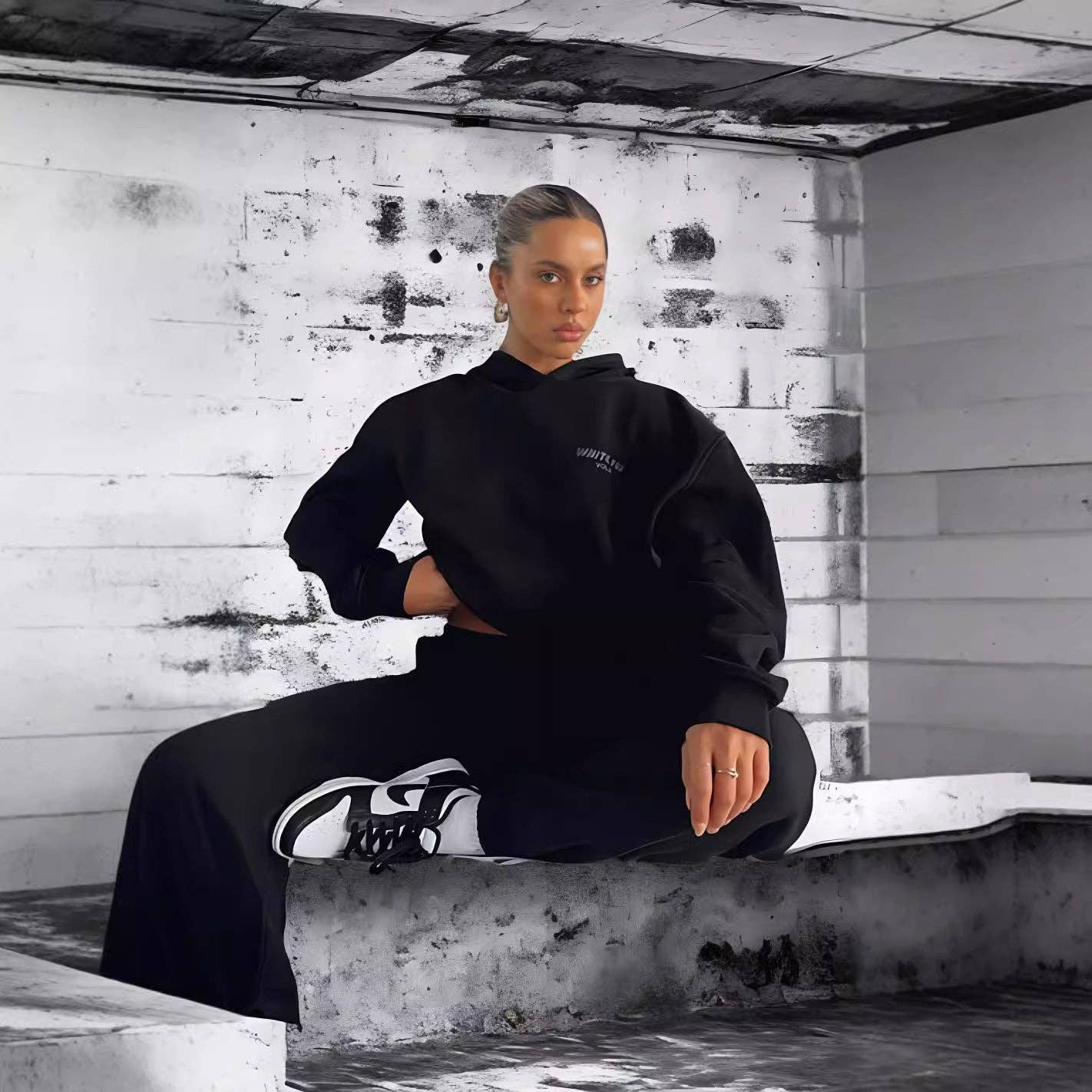 Person wearing a black hoodie and pants sitting on a concrete ledge with a distressed wall background.