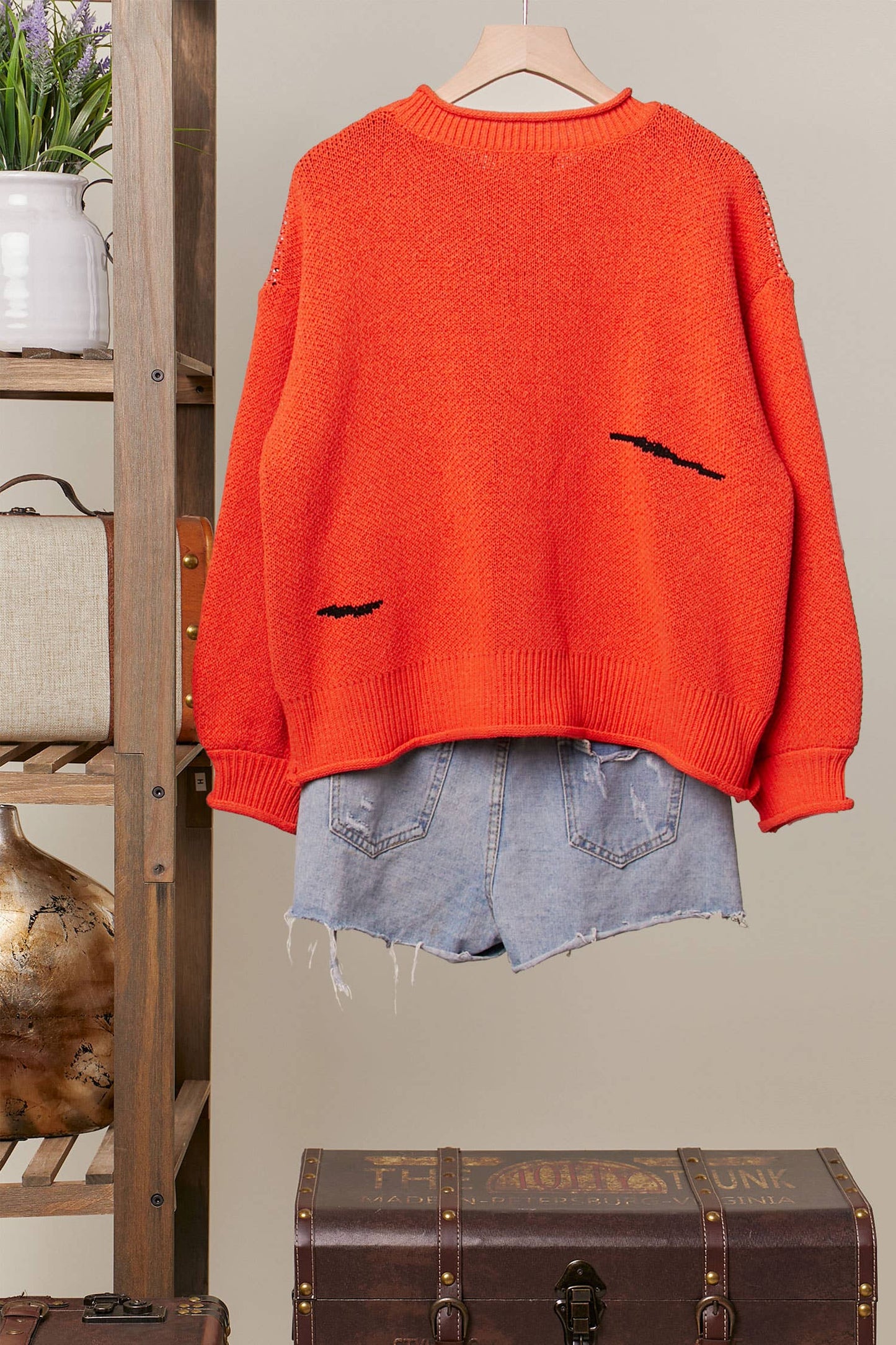 Orange sweater with black bat designs on a hanger against a neutral wall.