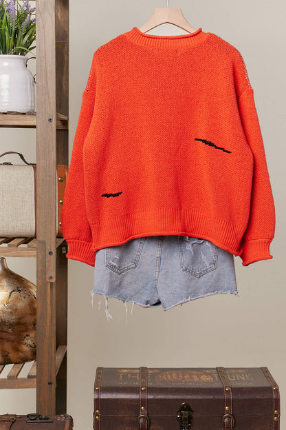 Orange sweater with black bat designs on a hanger against a neutral wall.