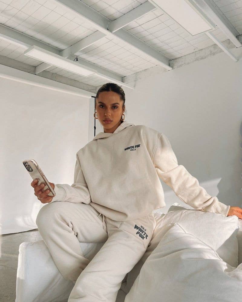Person wearing a beige hoodie and pants sitting on a white couch in a minimalistic room.