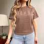 Person wearing a brown t-shirt with text and graphics, standing in a room with a lamp and plant.