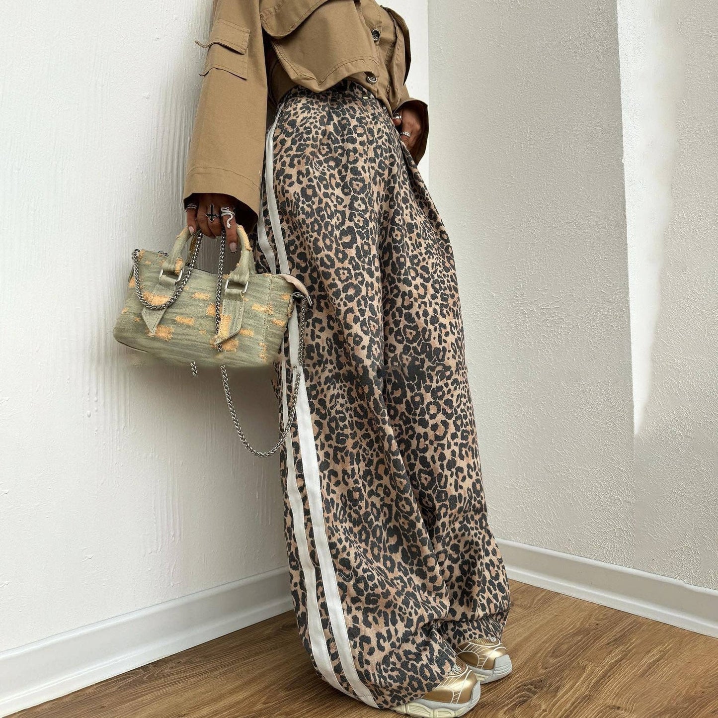 Person wearing leopard print pants with a beige jacket and holding a green handbag against a white wall.