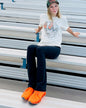 Person wearing a white t-shirt with a graphic design, dark pants, and orange shoes sitting on metal bleachers.