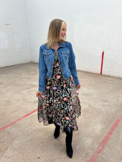 Woman wearing a floral dress and denim jacket in an indoor setting with a plain white wall.