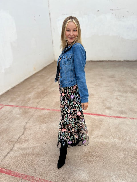 Woman wearing a denim jacket and floral dress standing in an indoor setting with a white wall and concrete floor.