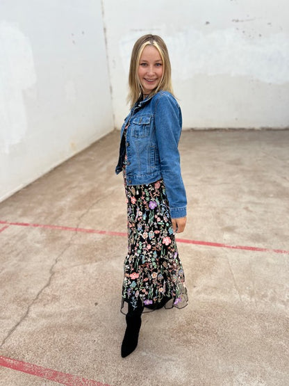Woman wearing a denim jacket and floral dress standing in an indoor setting with a white wall and concrete floor.