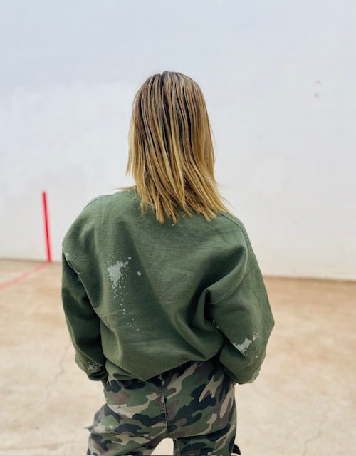 Person wearing a green sweatshirt with white paint splashes and camouflage pants in an indoor setting.