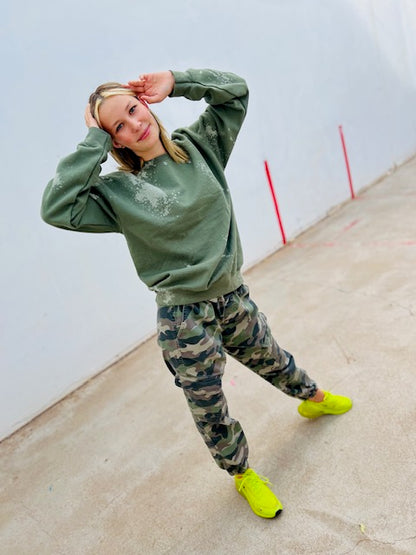 Person wearing a green sweatshirt and camouflage pants with bright yellow shoes on a plain background
