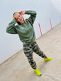 Person wearing a green sweatshirt and camouflage pants with bright yellow shoes on a plain background