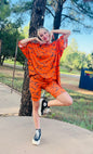 Person wearing an orange outfit with black patterns, standing on a sidewalk with trees and grass in the background.
