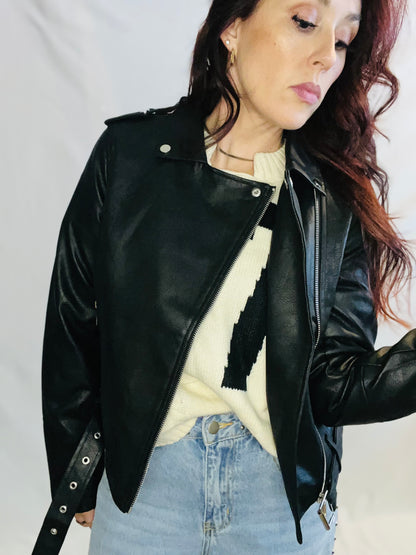 Person wearing a black leather jacket over a cream sweater and blue jeans on a plain background
