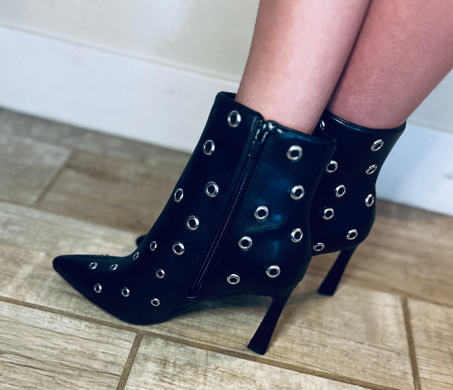 Black high-heeled boots with silver eyelets worn indoors on a tiled floor.