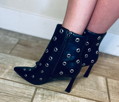 Black high-heeled boots with silver eyelets worn indoors on a tiled floor.