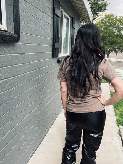 Person wearing a brown shirt and black leather pants standing on a sidewalk next to a gray house.