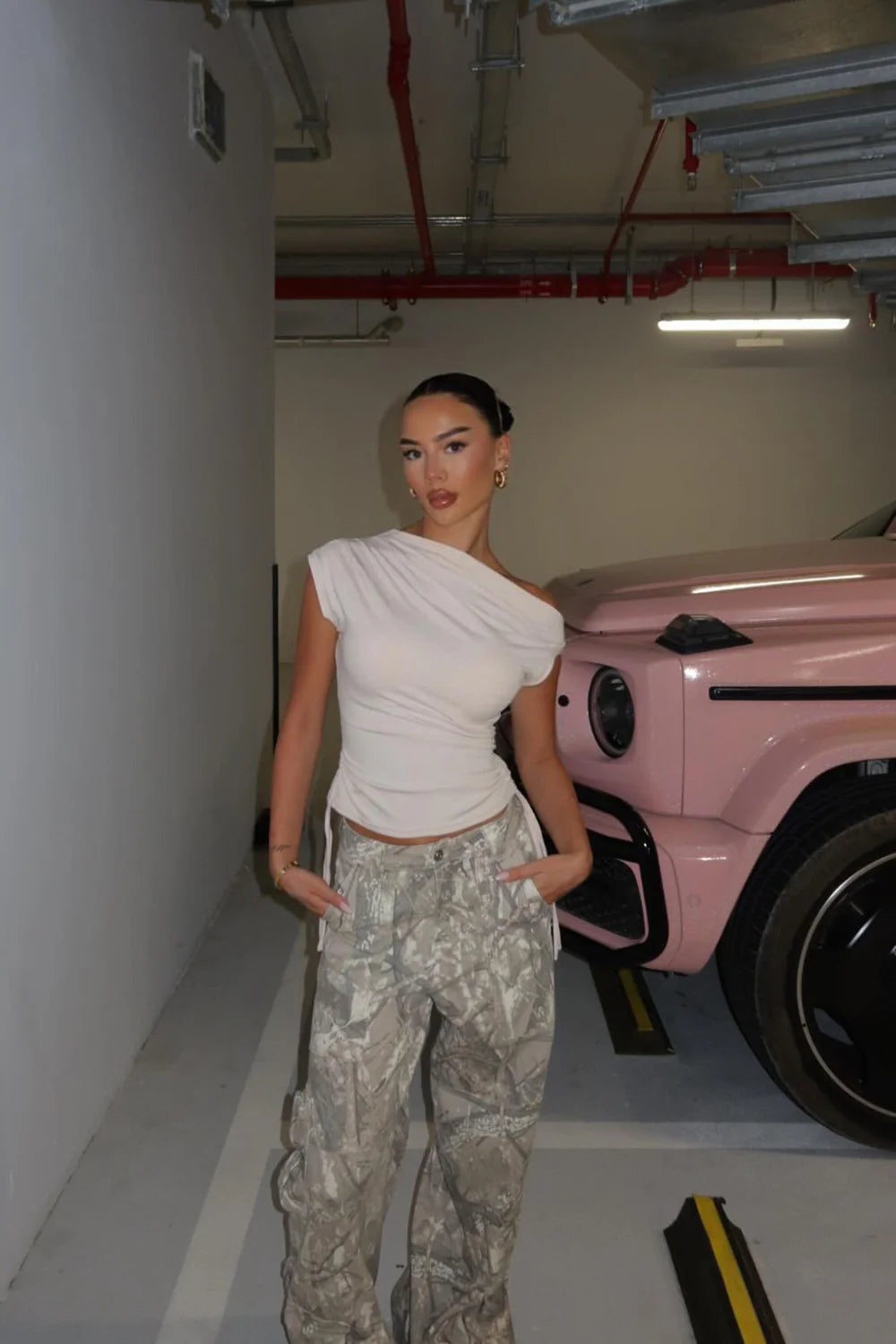Woman in a white top and camouflage pants standing in a garage with a pink car.