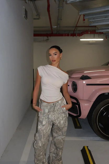 Woman in a white top and camouflage pants standing in a garage with a pink car.