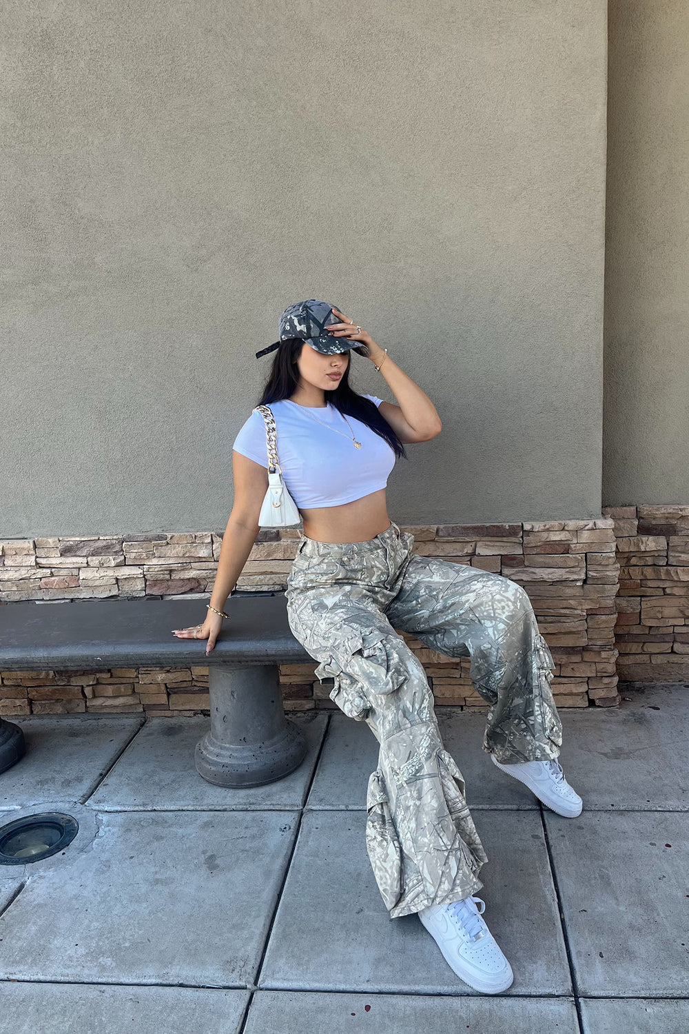 Woman sitting on a bench wearing a crop top, camouflage pants, and white sneakers.