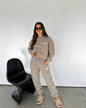 Woman in a beige tracksuit standing next to a black chair against a white wall