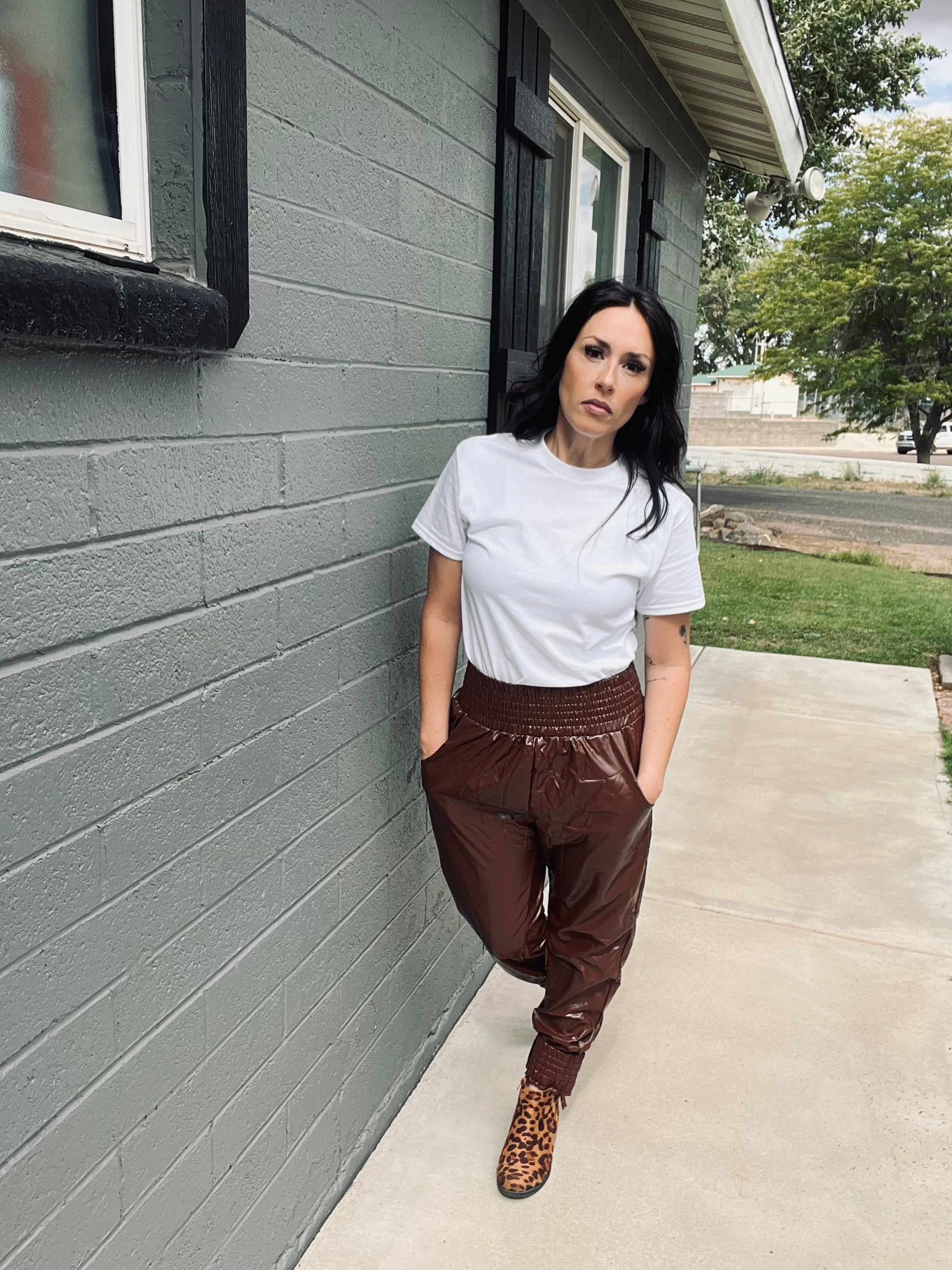 Woman in white t-shirt and brown pants standing against a gray brick wall.
