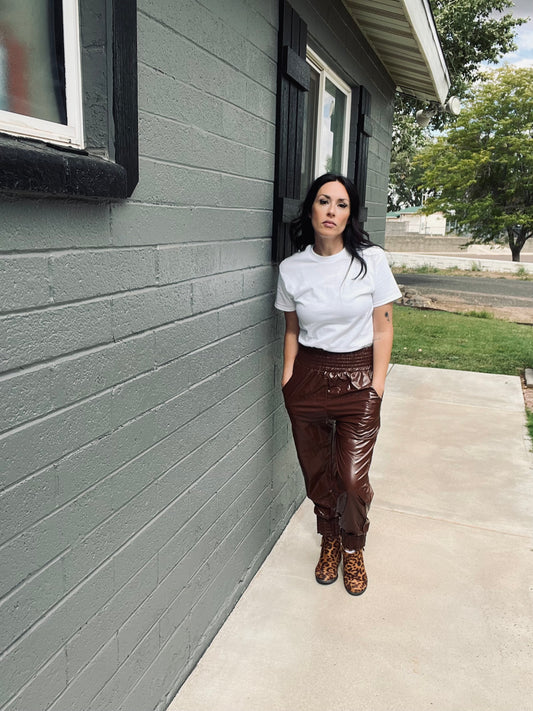 Woman standing against a gray house wall wearing a white t-shirt and brown pants.