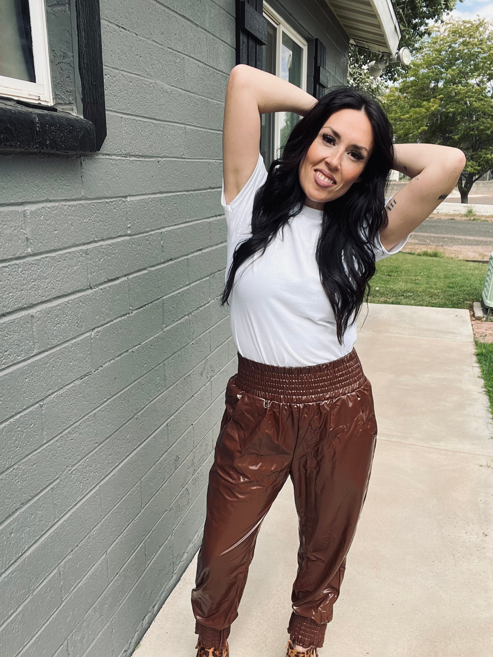 Woman posing against a gray wall wearing a white top and brown pants.