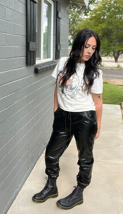 Person wearing a white graphic t-shirt, black leather pants, and black boots standing against a gray brick wall.