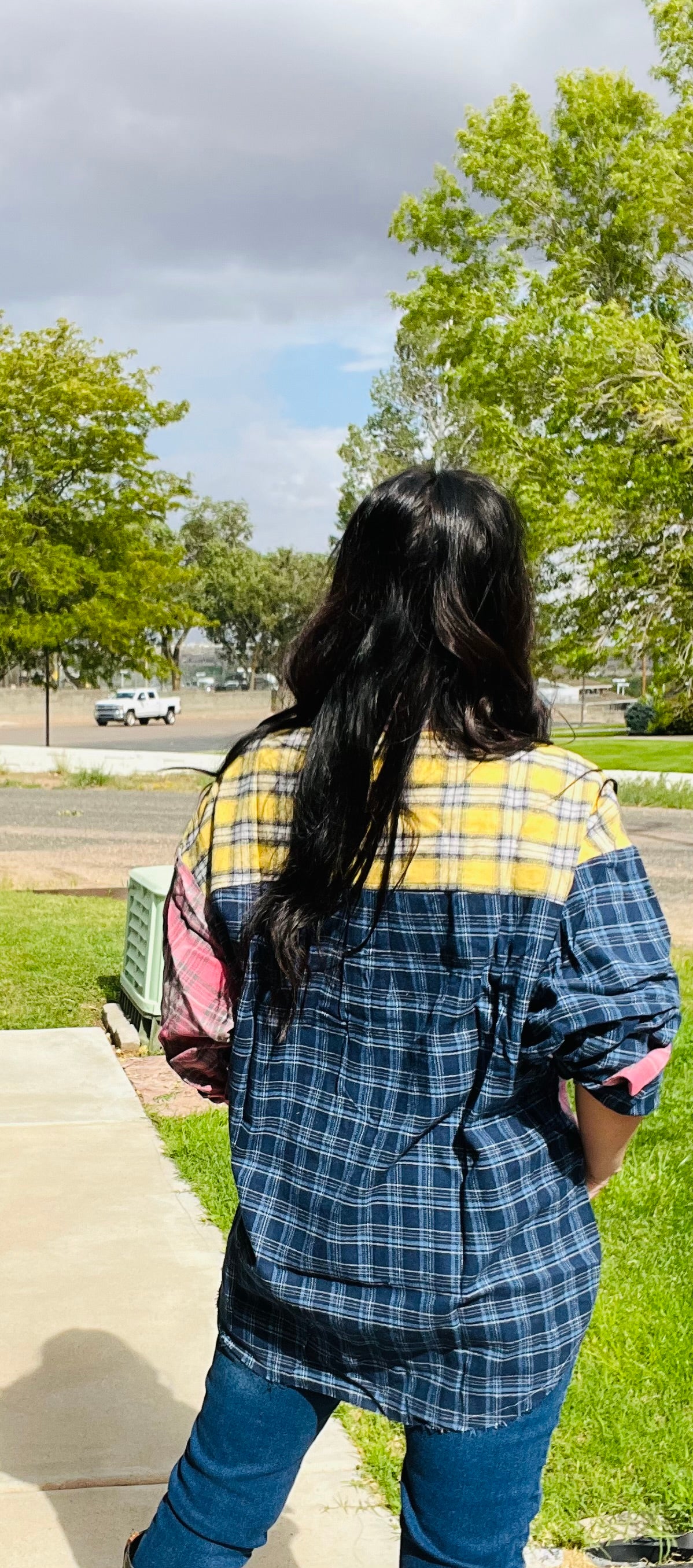 Person wearing a blue and yellow plaid shirt walking on a sidewalk with trees and cars in the background.