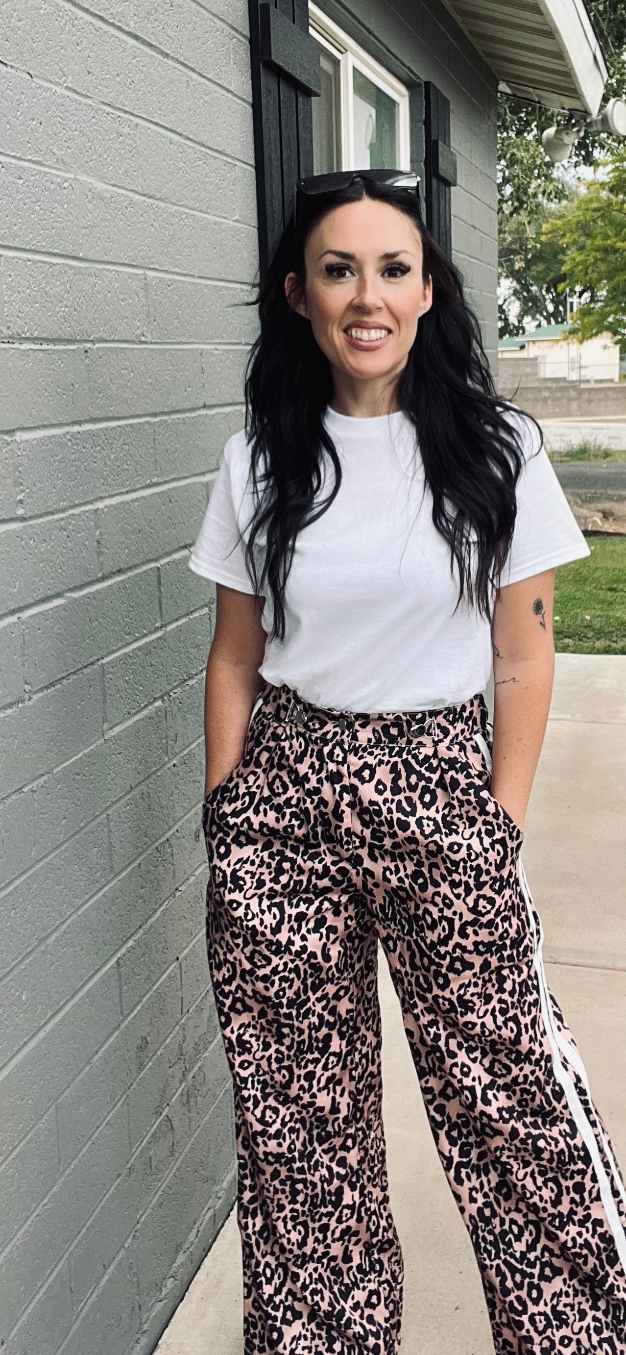 Woman wearing a white t-shirt and leopard print pants standing against a gray wall.