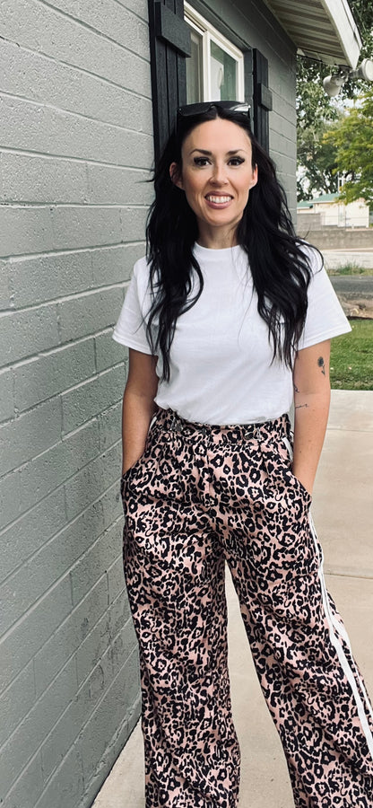 Woman wearing a white t-shirt and leopard print pants standing against a gray wall.