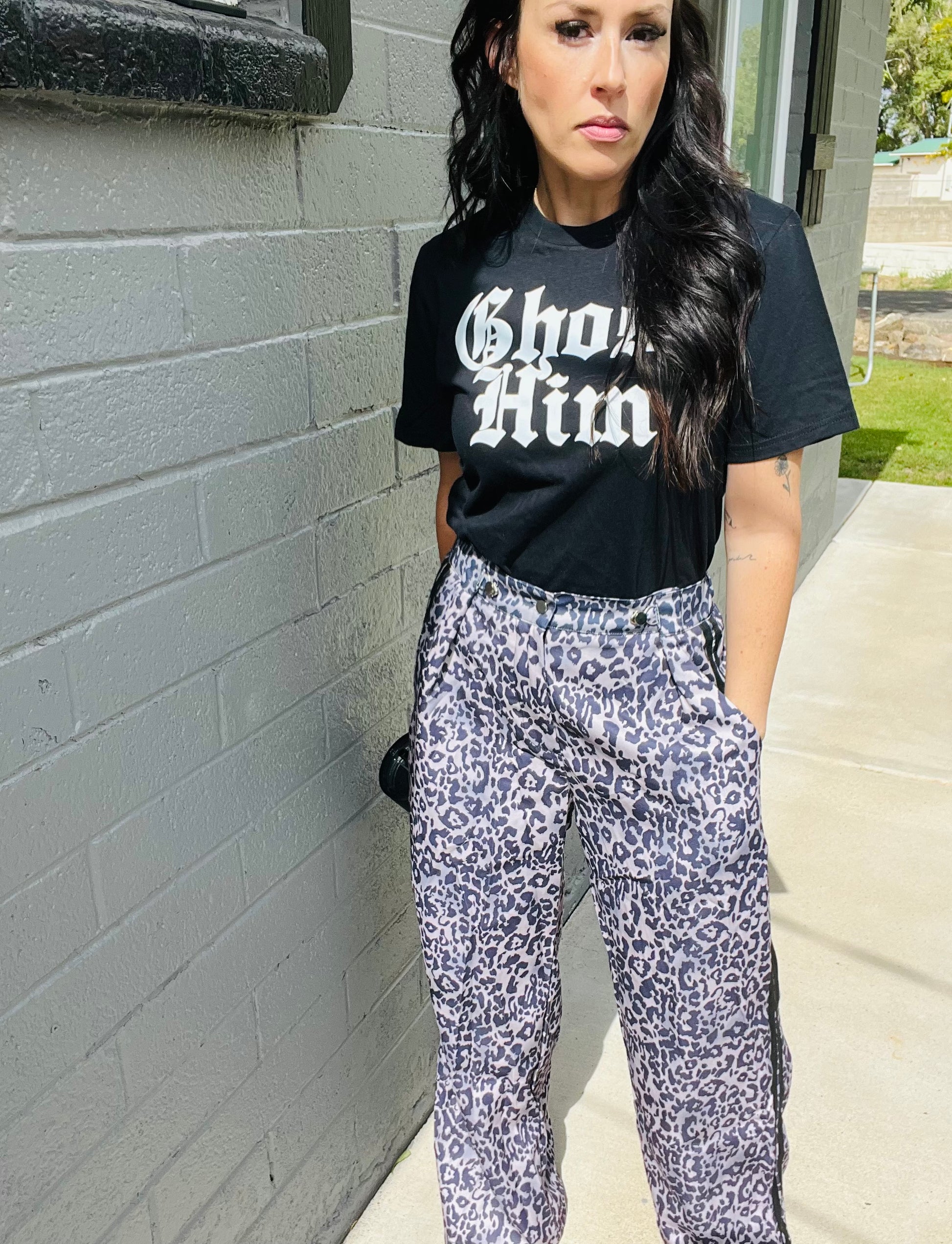 Woman wearing a black t-shirt with white text and patterned pants standing against a gray wall.