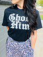 Person wearing a black t-shirt with white text and patterned pants standing against a gray wall.