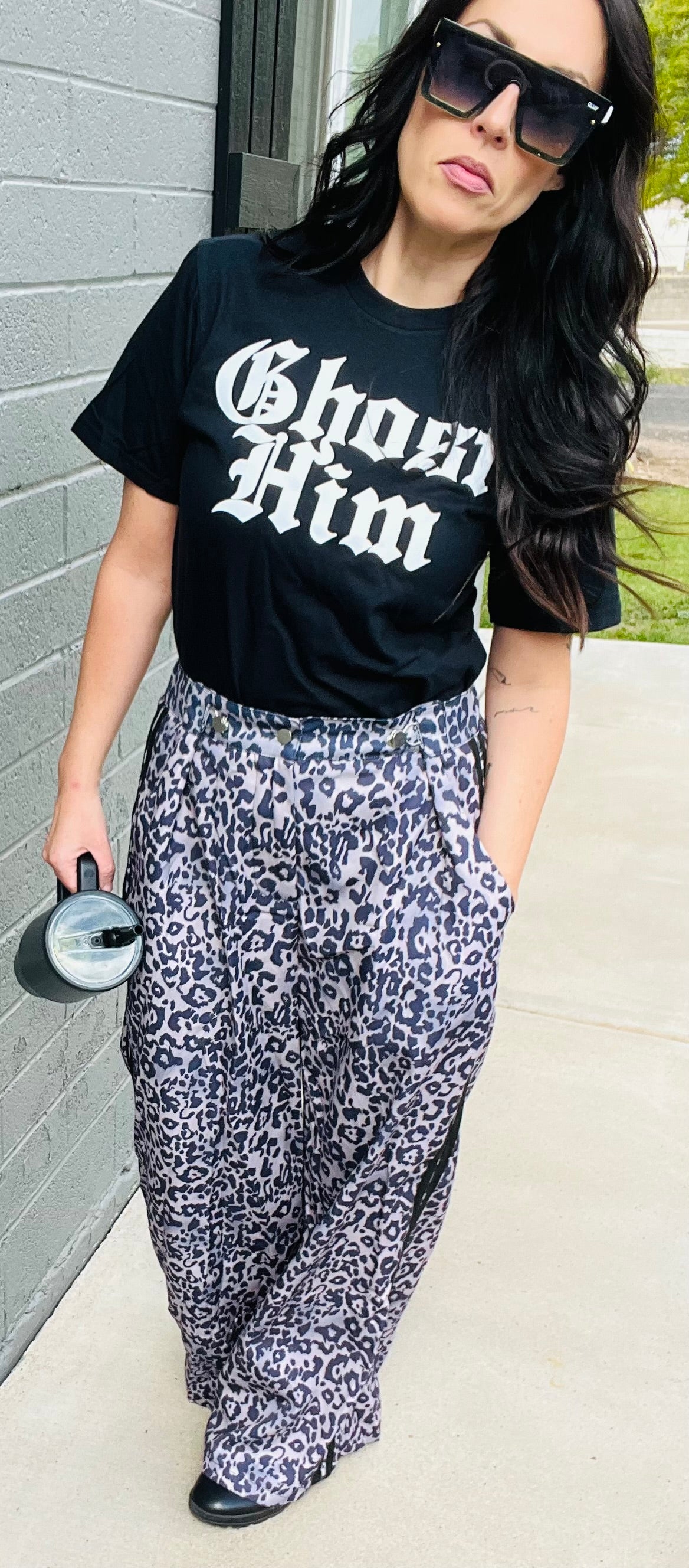 Woman wearing a black t-shirt with 'Ghost Him' text and patterned pants against a gray wall.