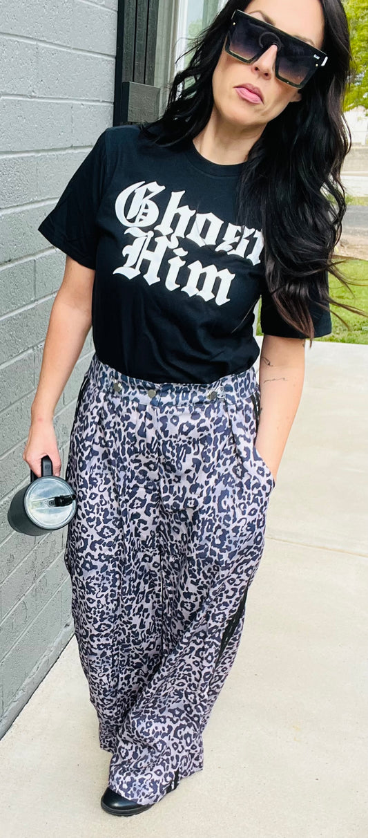 Woman wearing a black t-shirt with 'Ghost Him' text and patterned pants against a gray wall.