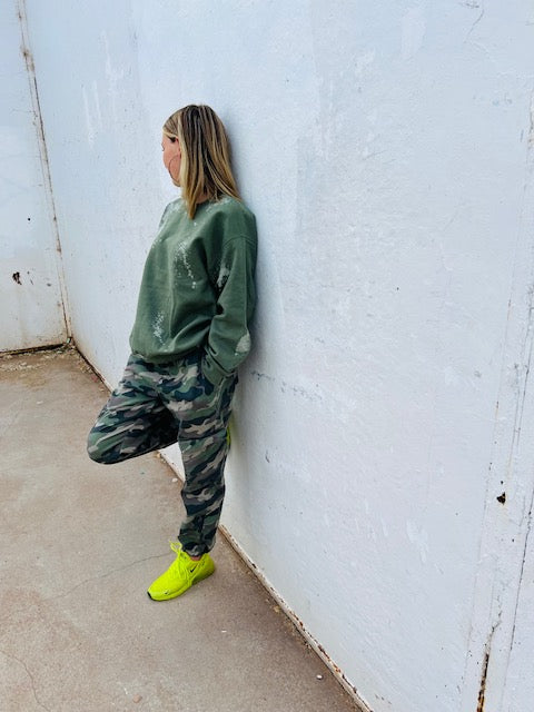Person wearing a green sweatshirt and camouflage pants leaning against a white wall.