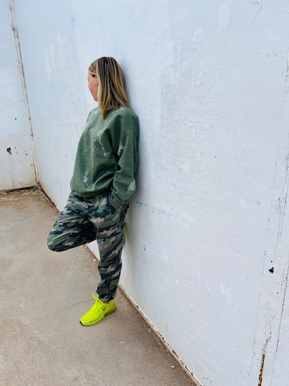Person wearing a green sweatshirt and camouflage pants leaning against a white wall.
