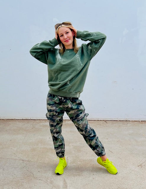 Person wearing a green sweatshirt and camouflage pants with neon green shoes against a light blue wall.