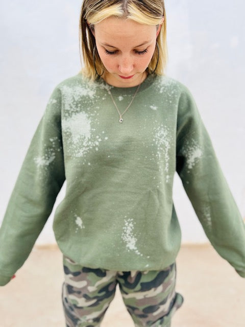 Person wearing a green sweatshirt with white splatter design and camouflage pants on a plain background