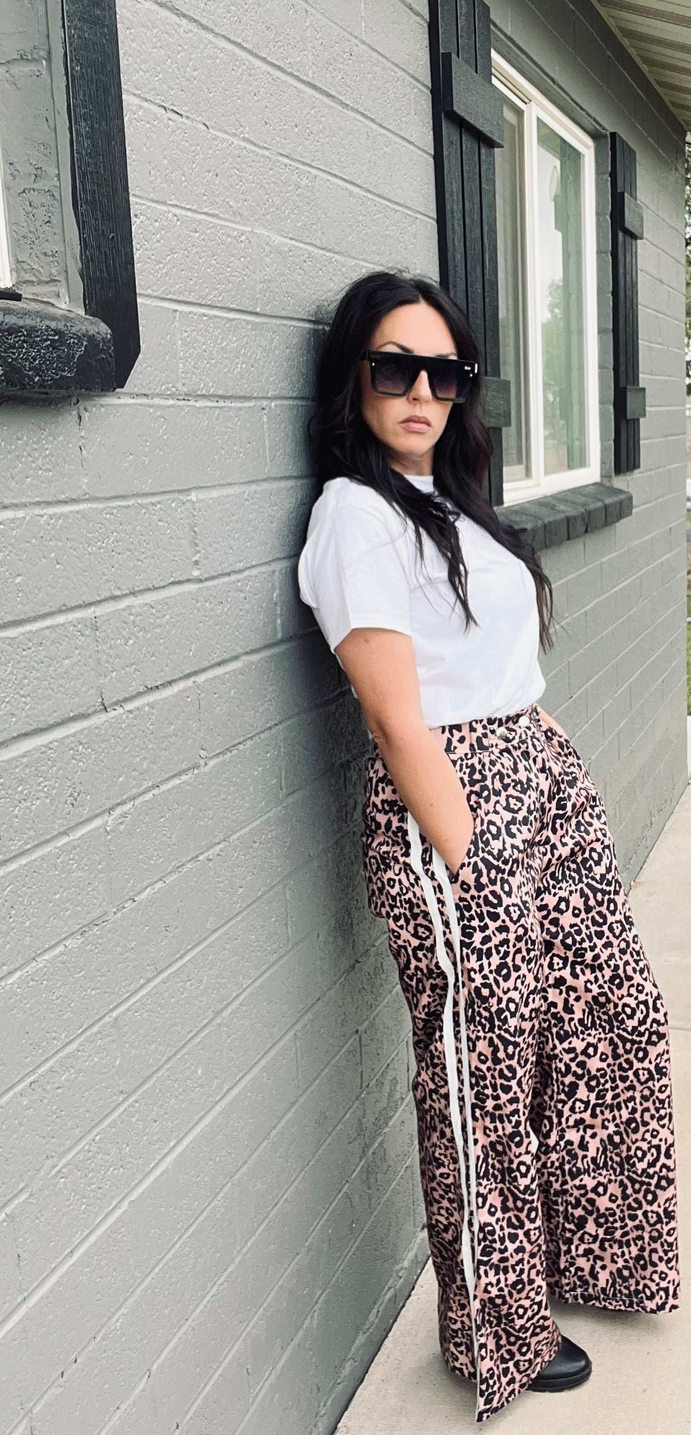 Woman wearing a white shirt and leopard print pants leaning against a gray brick wall.