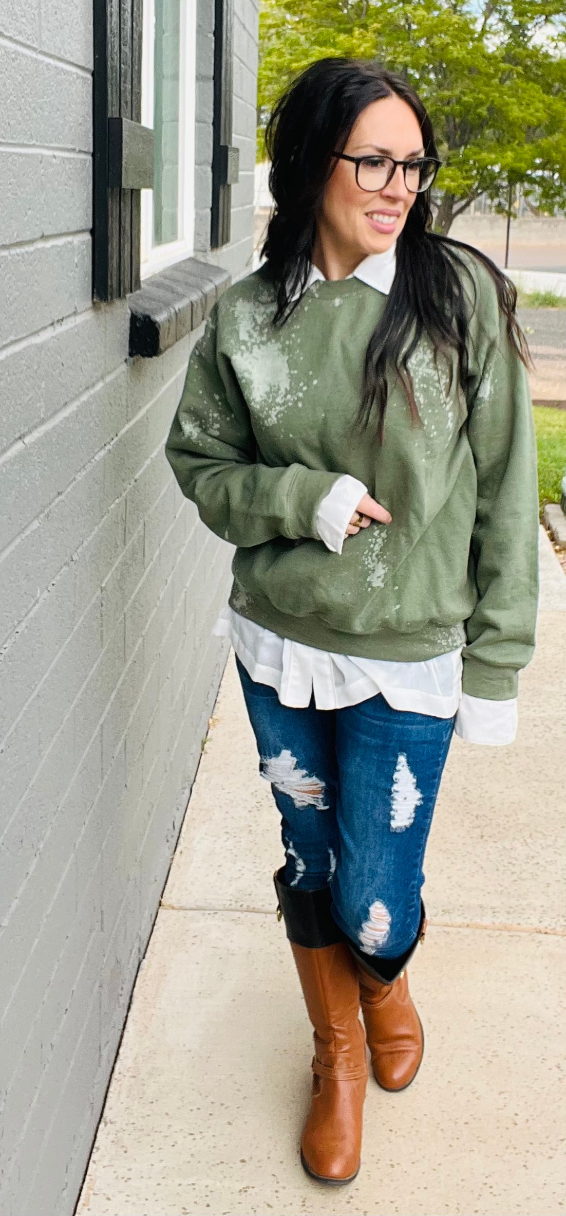 Woman wearing a green distressed sweater, white shirt, blue jeans, and brown boots standing against a gray wall.