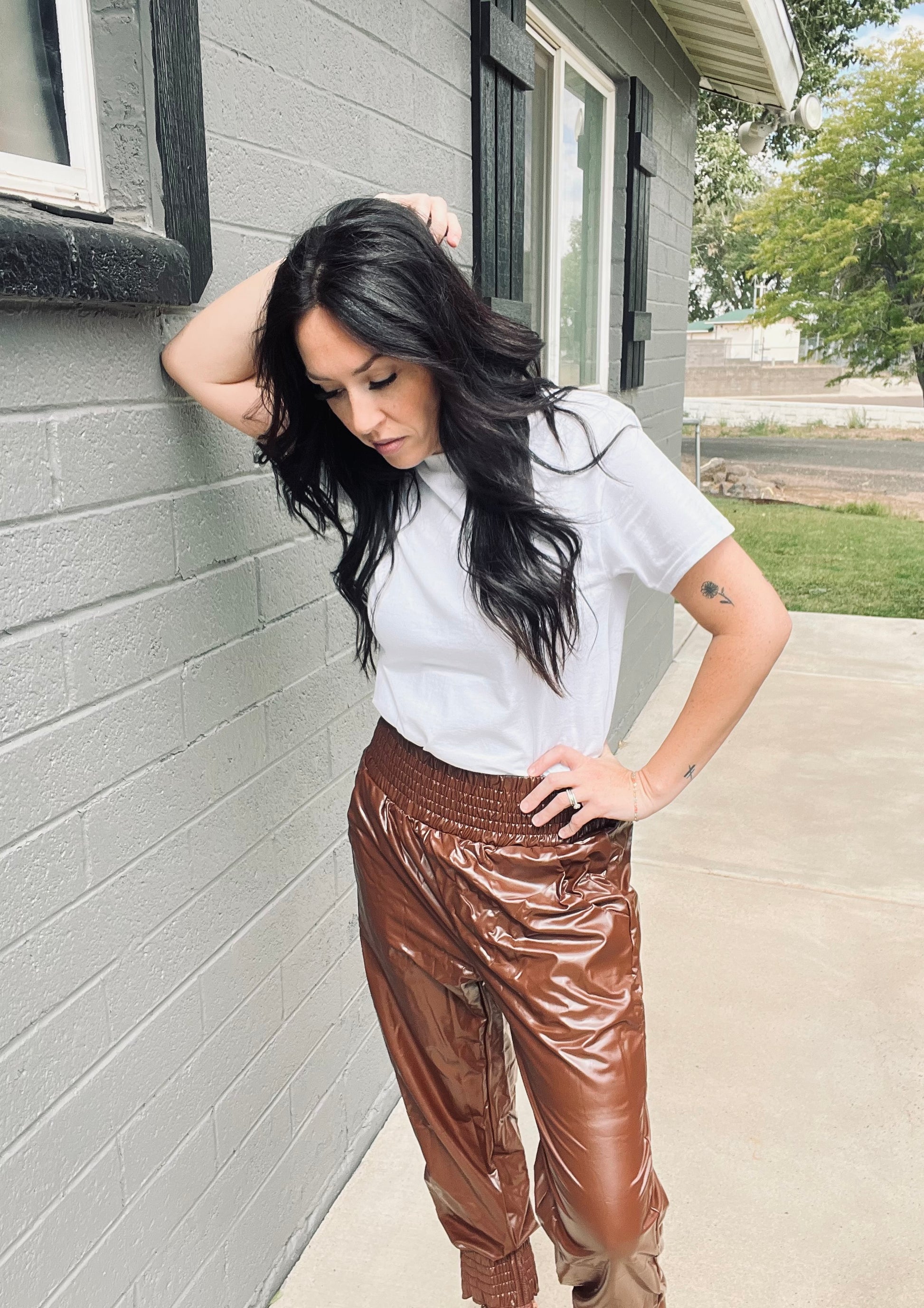 Woman wearing a white shirt and brown shiny pants standing against a gray brick wall.