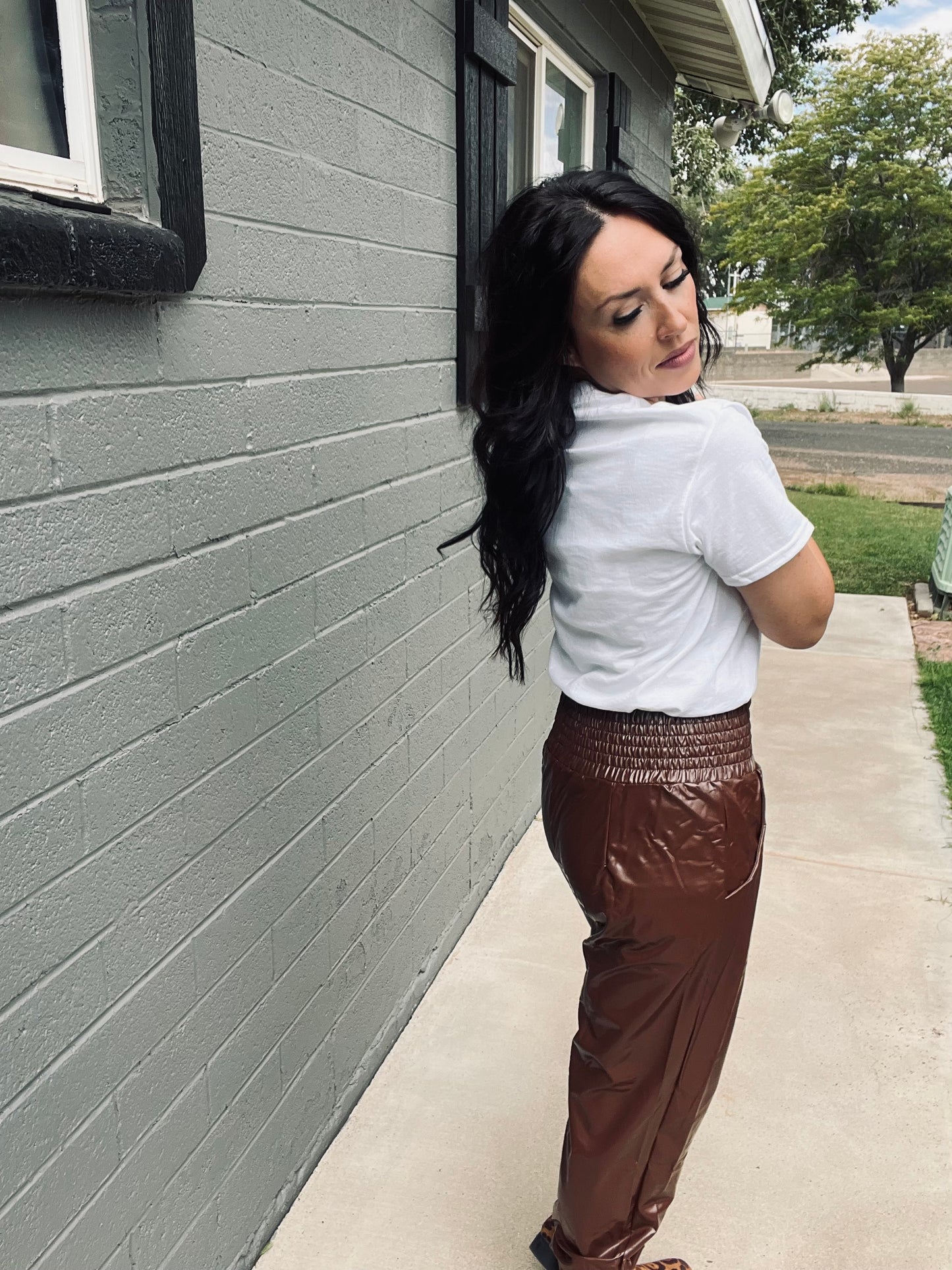 Woman in a white shirt and brown pants leaning against a gray wall.