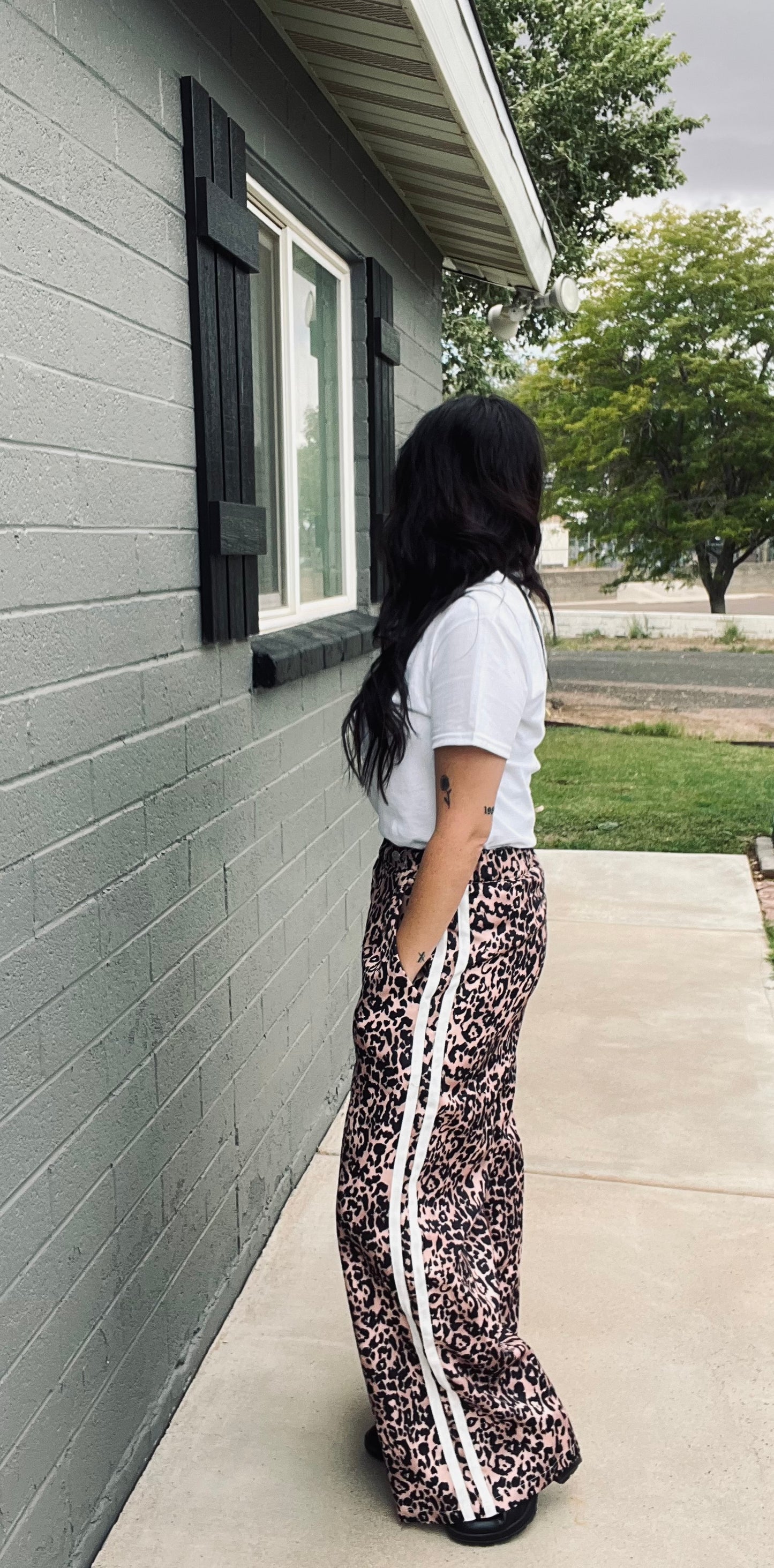 Person wearing a white t-shirt and patterned pants standing against a gray house exterior.