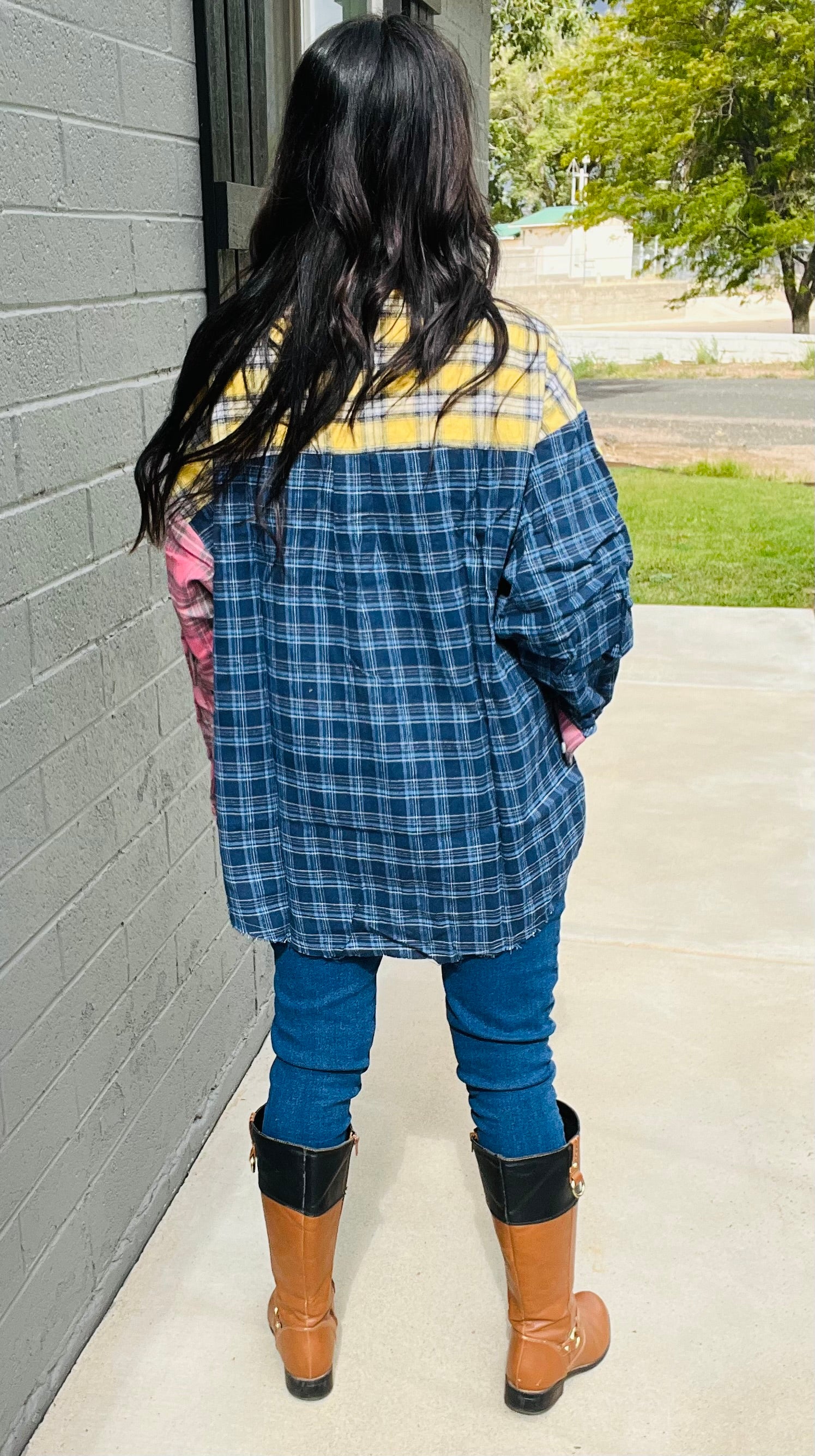 Person wearing a blue plaid jacket with yellow accents, blue jeans, and brown boots standing against a gray wall.